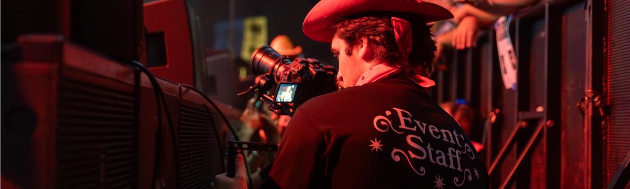 A student wearing a cowboy hat and a t-shit that says "Event Staff" filming at a concert